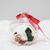 Boule de Noël en verre ou est endormi un bébé fille habillée de blanc et vert avec son chapeau blanc et son ours brun