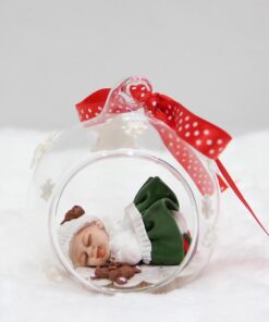 Boule de Noël en verre ou est endormi un bébé fille habillée de blanc et vert avec son chapeau blanc et son ours brun